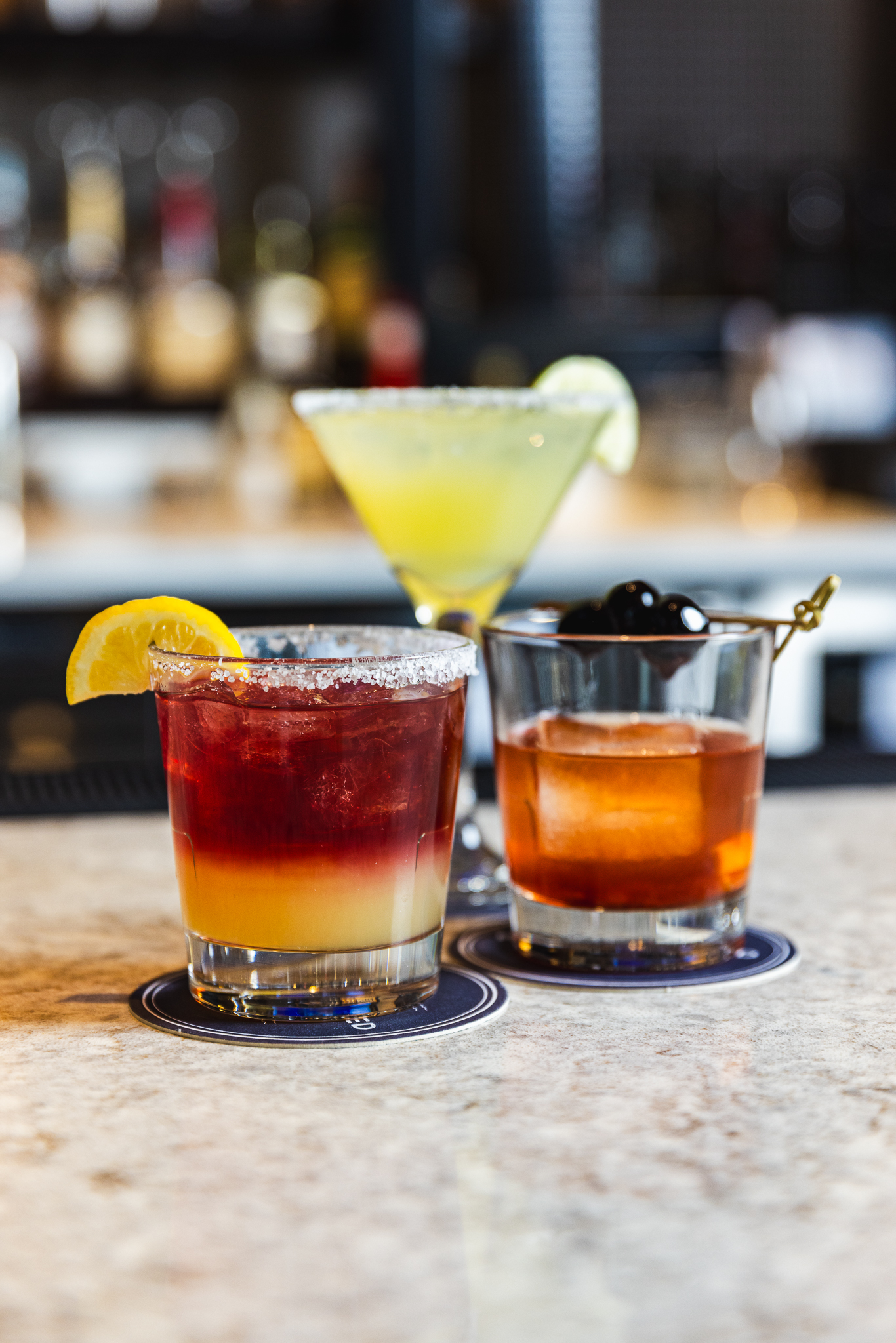 Three craft cocktails on the bar at The Placery — a layered red and orange cocktail, a margarita, and an old fashioned with brandied cherries