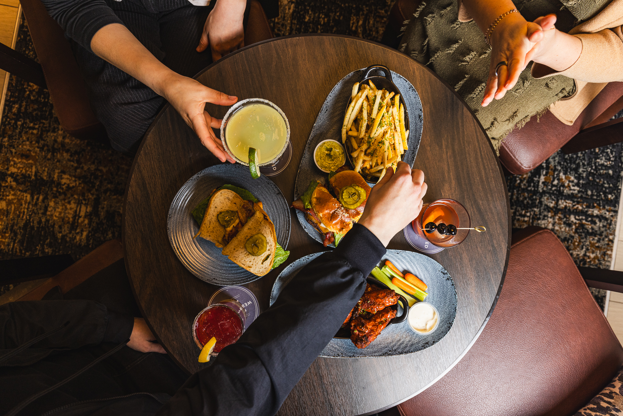 Overhead shot of a Placery table spread with a bacon chicken sandwich, smash burger and fries, buffalo wings with carrots and celery, and craft cocktails inside the Topgolf Swing Suite