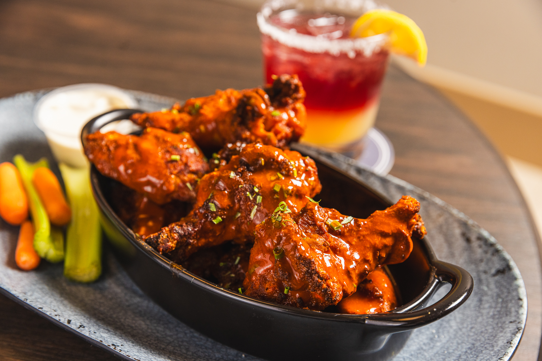 Bowl of buffalo chicken wings with celery, carrots, and ranch dipping sauce alongside a red layered cocktail at The Placery