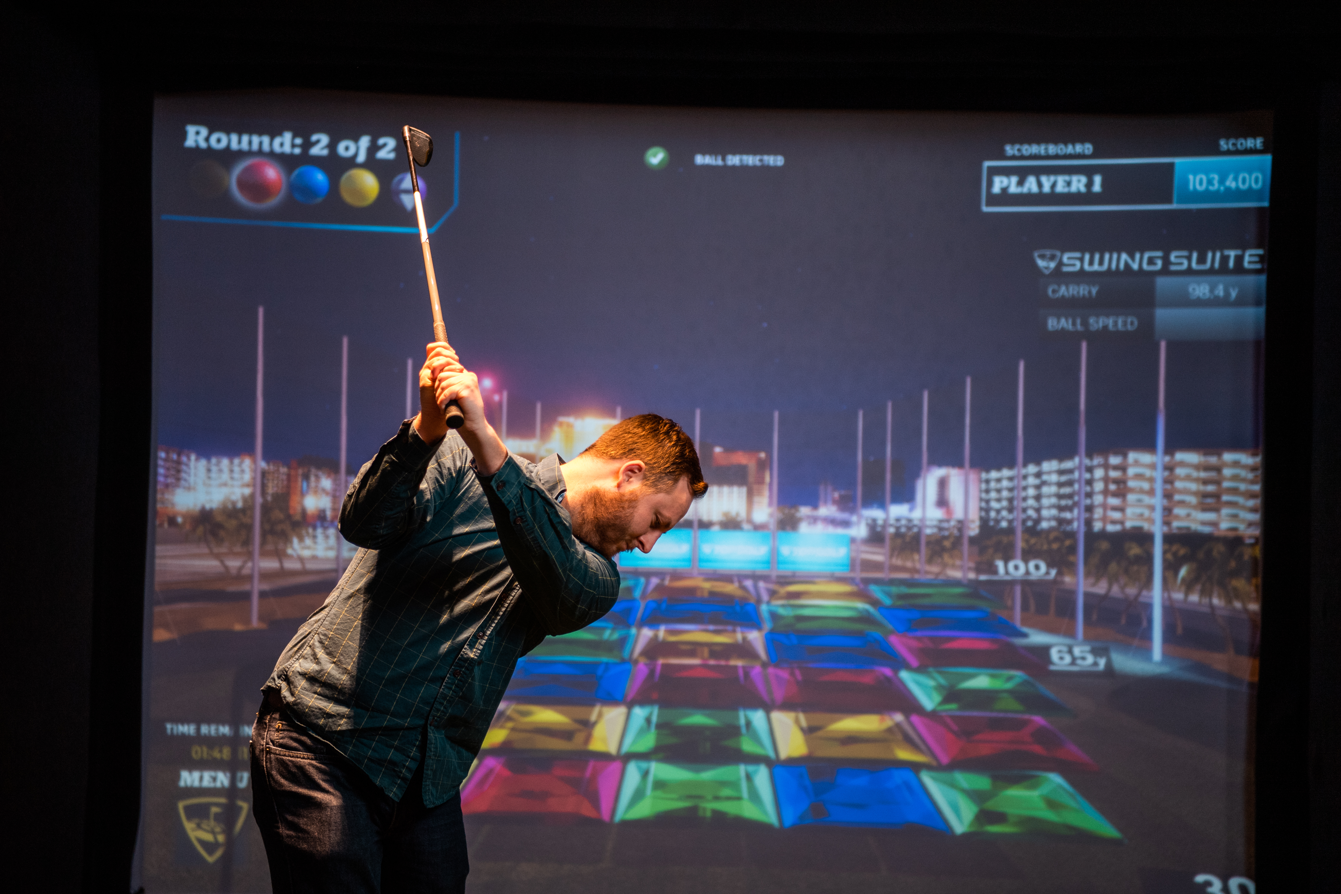 Player mid-swing in front of a colorful Gem Match game on the Topgolf Swing Suite screen