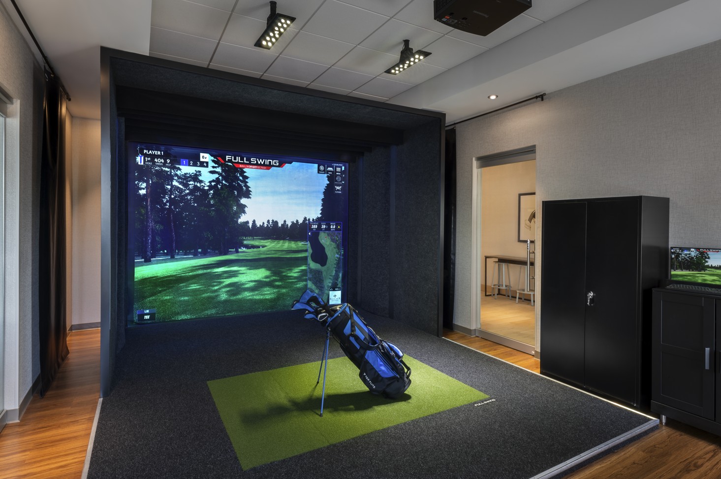 The actual Topgolf Swing Suite at Hyatt Place Munster — Full Swing simulator screen displaying a wooded golf course, golf bag on the artificial turf hitting mat, and entrance to the bay